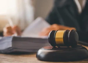 a wooden judge's gavel on a desk with a blurred defense lawyer reviewing a stack of court documents for a drug case trial in texas.