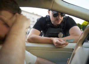 law enforcement officer reviewing a driver's license at the window of a car while a stressed driver holds his head during a traffic stop.